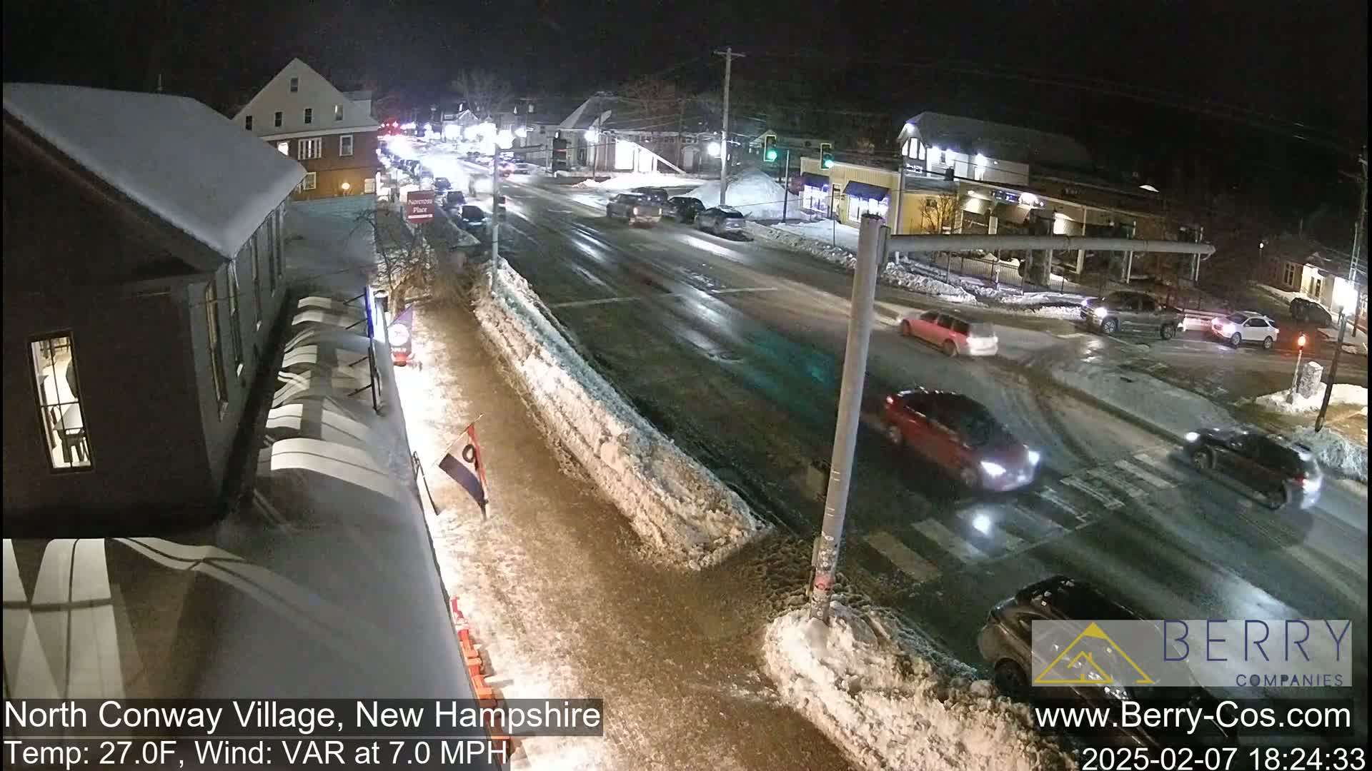 A snow-covered street scene at night shows several cars driving through a village intersection under a dark sky.