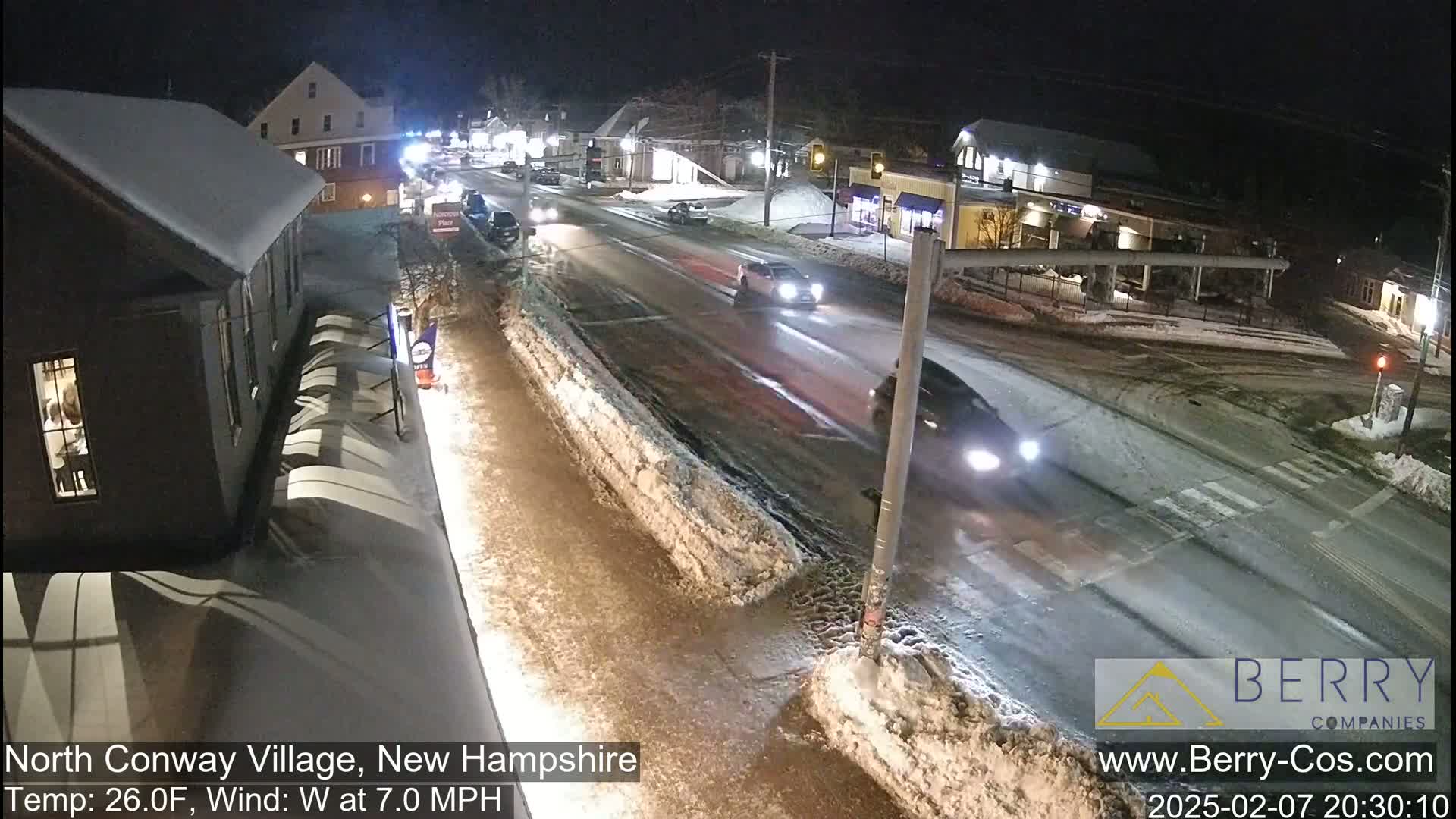 A snow-covered street intersection at night with several cars driving by under partly cloudy conditions.