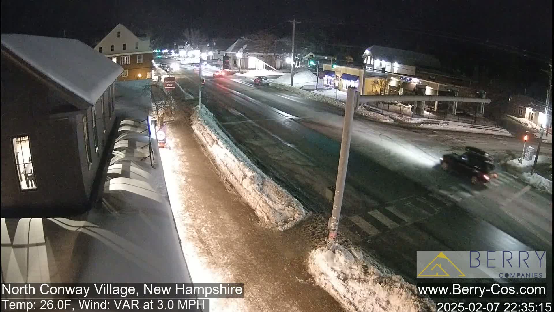 A snow-covered street scene at night shows a few vehicles driving past buildings with some snow on the ground and a temperature of 26 degrees Fahrenheit.
