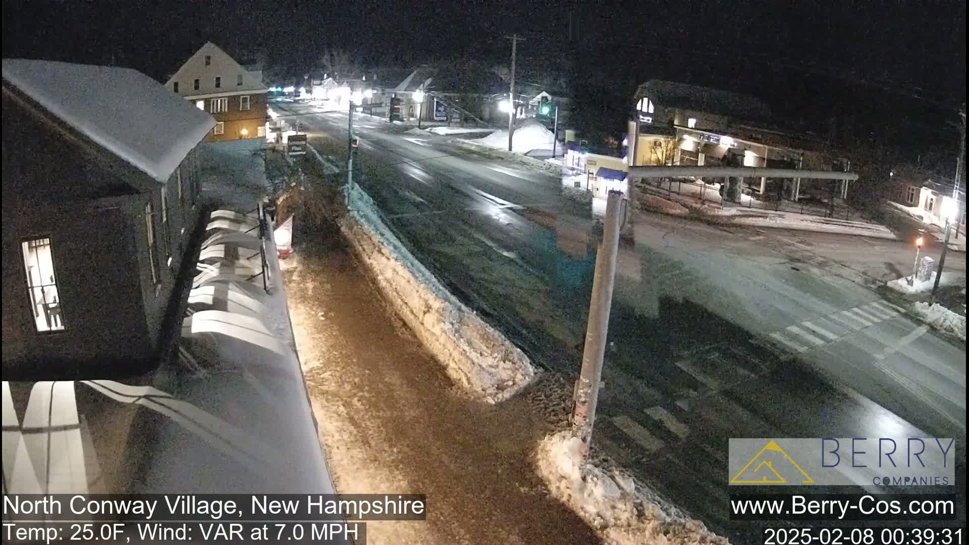 A nighttime view of a snow-covered street in a village, with buildings and streetlights visible under a dark sky.