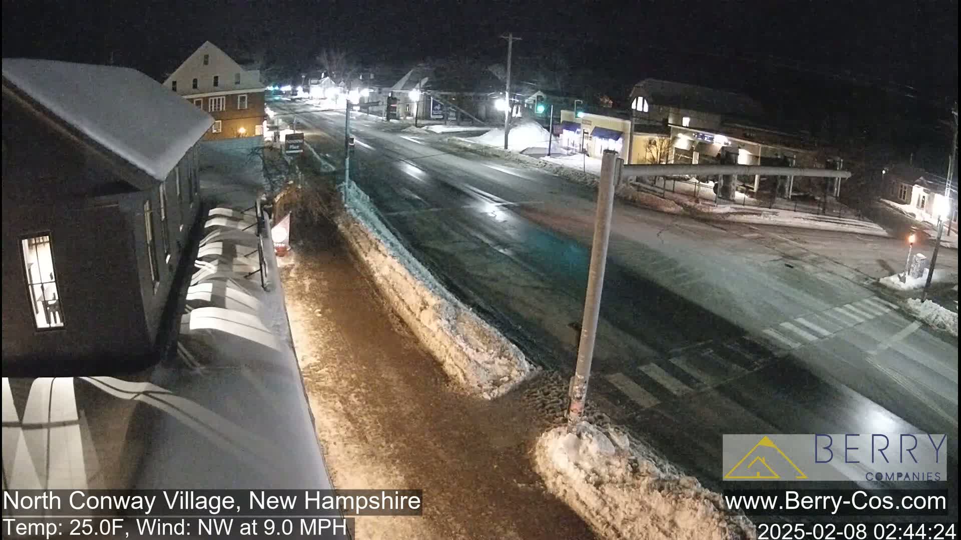 A snow-covered street scene at night shows a road intersection with buildings and some cars, under a clear sky.