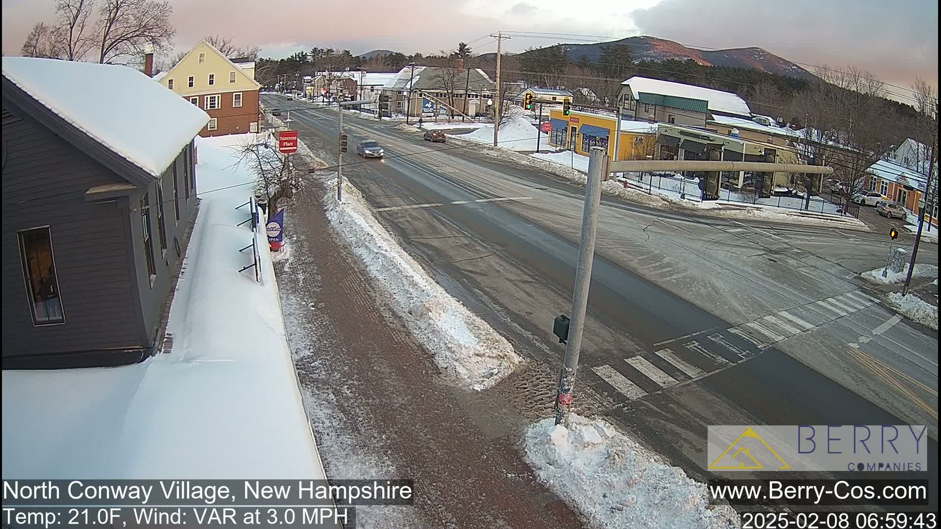 A snow-covered street scene in a small town shows a road intersection with several cars, buildings, and a partly cloudy sky.