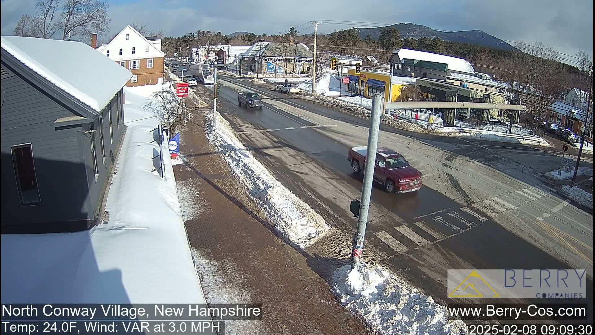 A partly sunny, winter day shows a road intersection in a small town with snow on the sides of the road and some melting snow and slush on the pavement; several vehicles are visible.