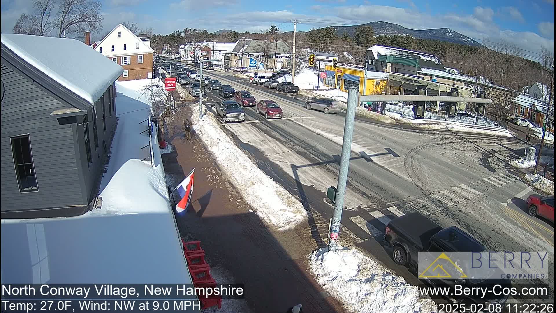 A sunny day in a snow-covered town shows a busy intersection with cars and pedestrians, and mountains in the distance.