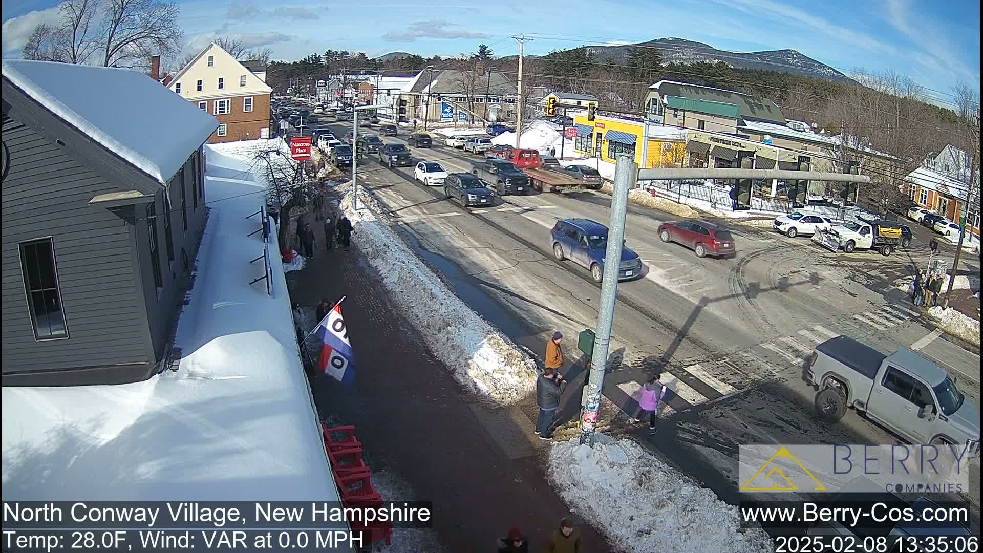 A sunny winter day in a village with snow-covered sidewalks and roads, showing several cars and pedestrians near a crosswalk.