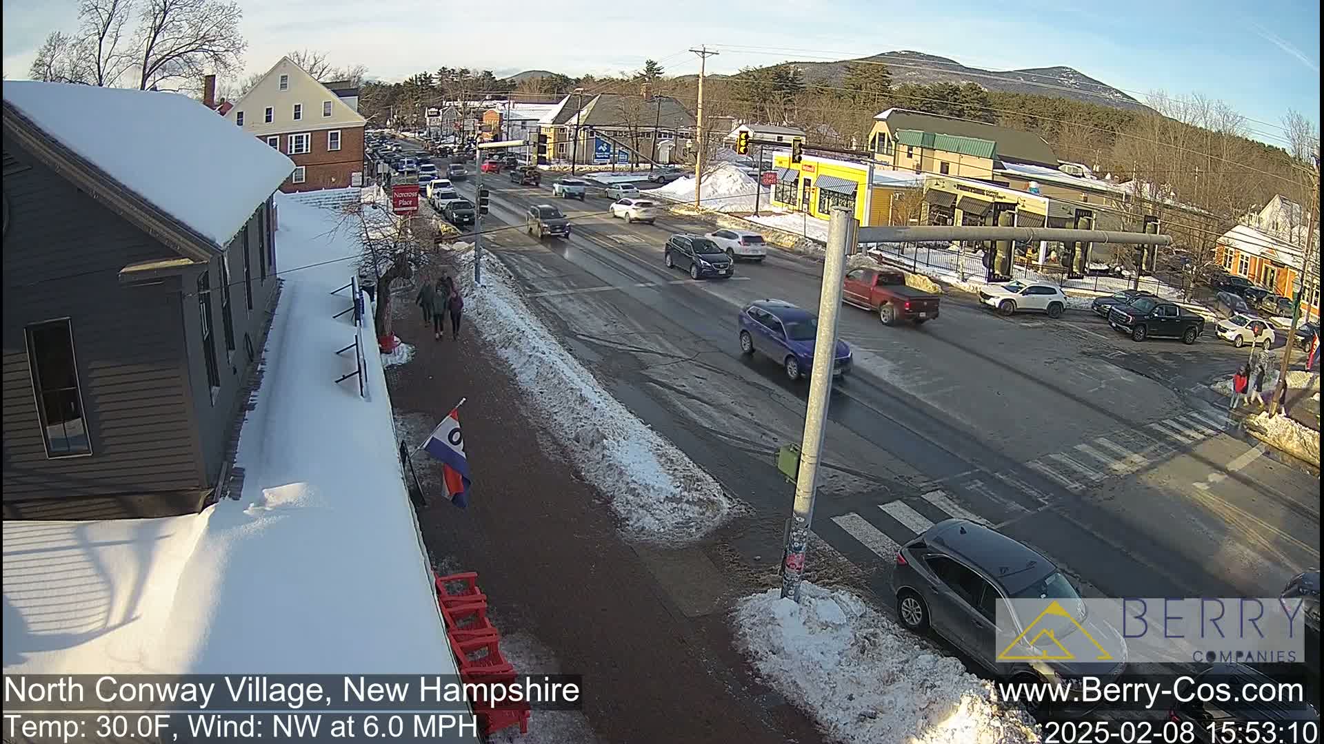 A snow-covered street scene shows cars driving on a road that intersects with a crosswalk, with pedestrians walking nearby under a sunny sky.
