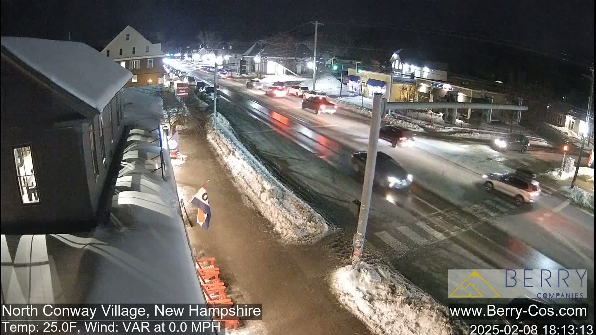 A nighttime street scene shows several cars driving on a wet road partially covered with snow, with buildings and snow-covered sidewalks lining the street.