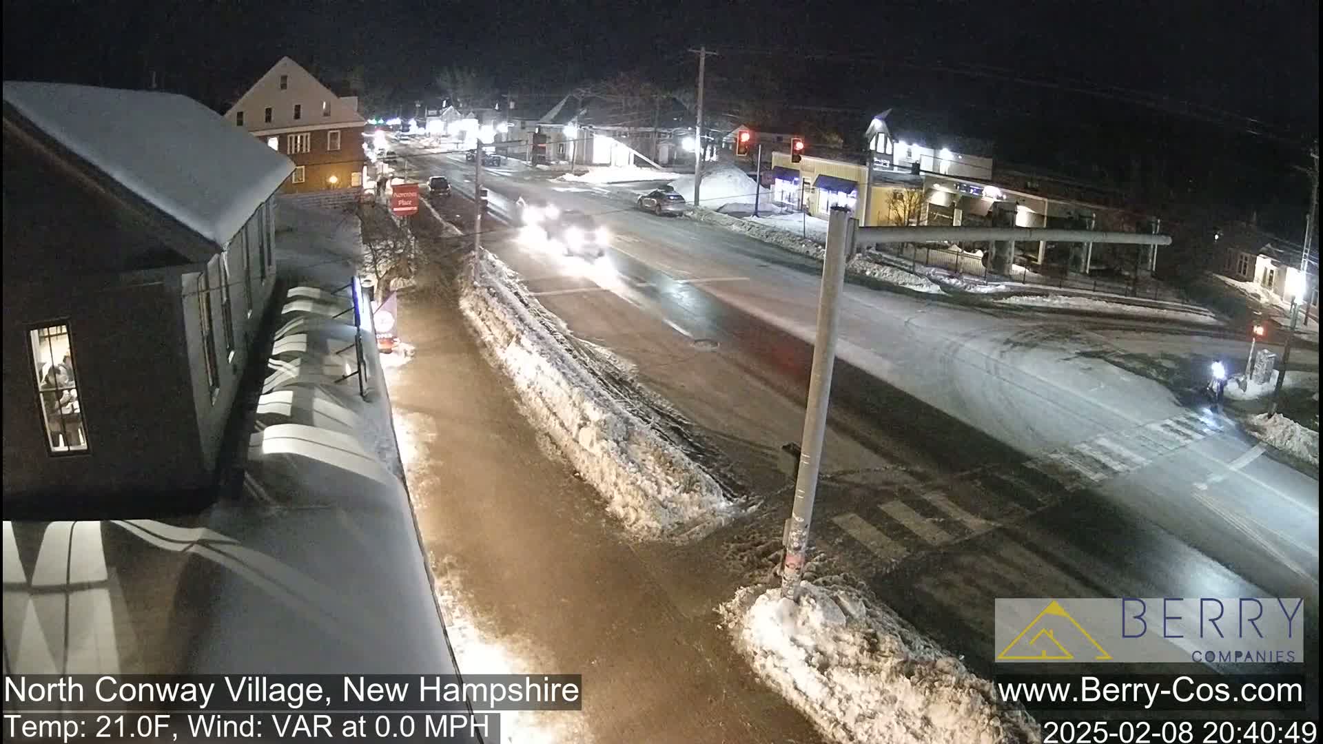 A nighttime street scene shows a few cars driving on a snow-covered road, with buildings and snow-covered sidewalks visible under a dark sky.