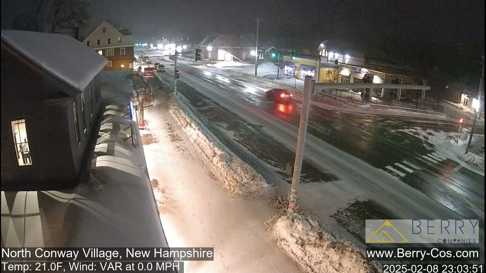 A nighttime street scene shows a snow-covered road with a few cars driving on it, under a dark sky.
