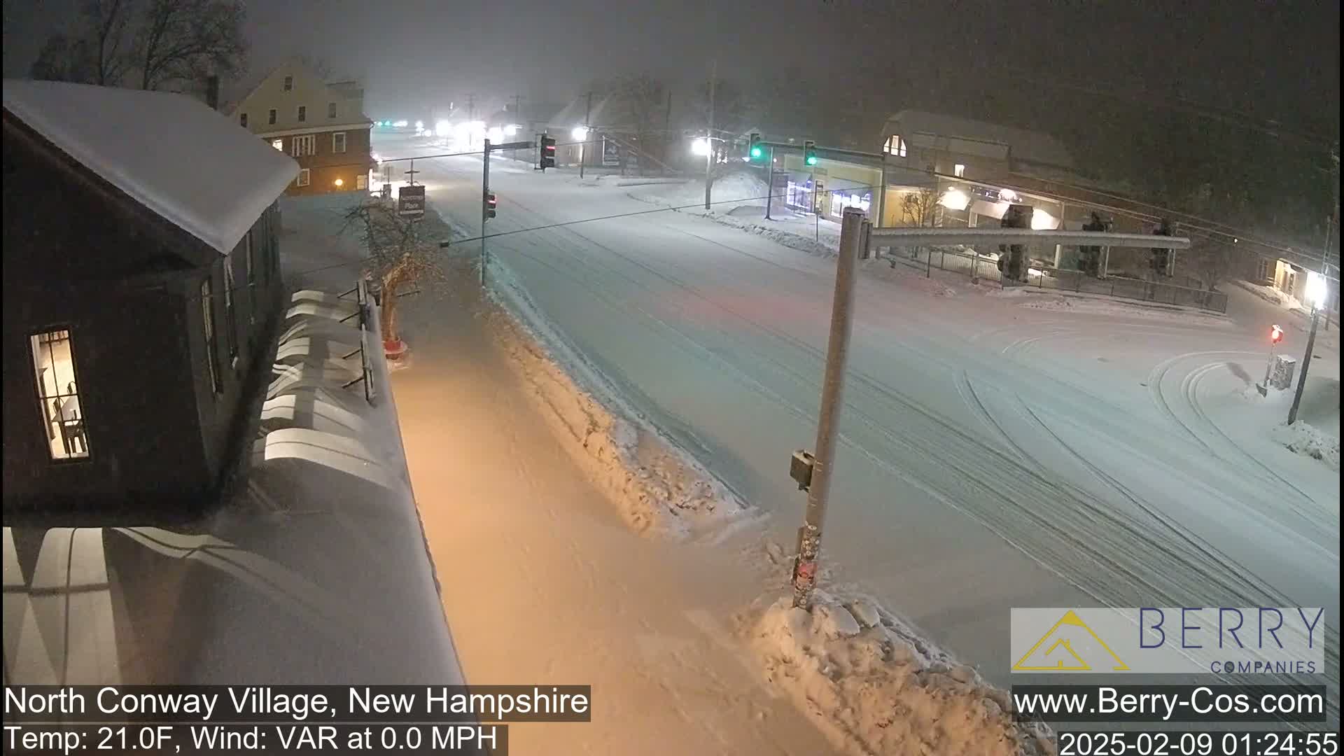 A snow-covered street scene at night shows a sparsely trafficked intersection with buildings covered in snow.