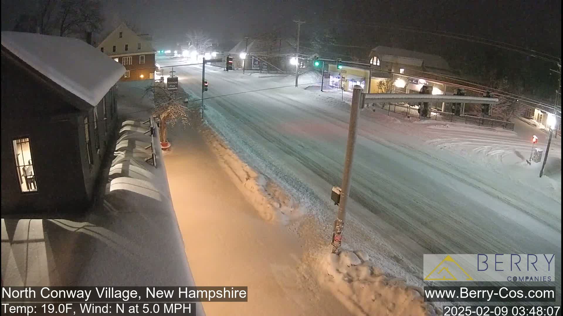 A snow-covered street scene at night shows a sparsely trafficked road, buildings covered in snow, and lightly falling snow with a north wind.