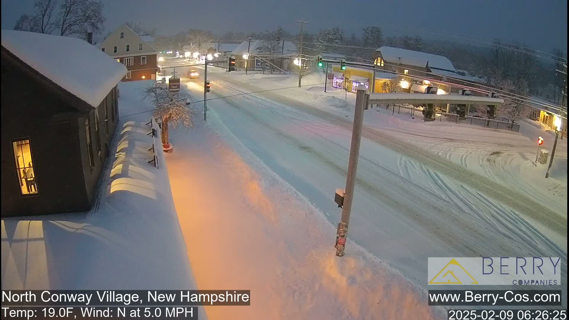 A snow-covered street scene at twilight shows cars driving on a road lined with snow-covered buildings under a dark sky.