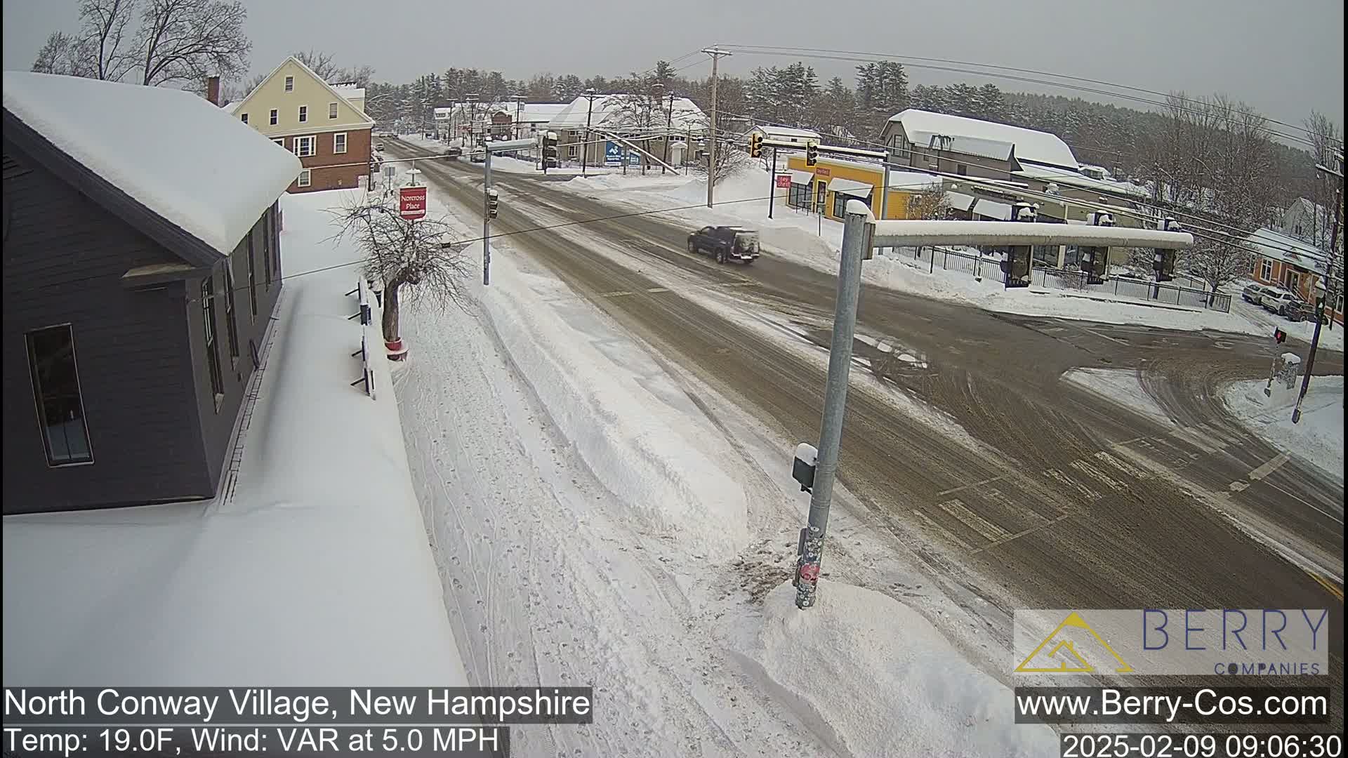 A snow-covered street scene shows a road intersection with several buildings and a few vehicles, under overcast skies.
