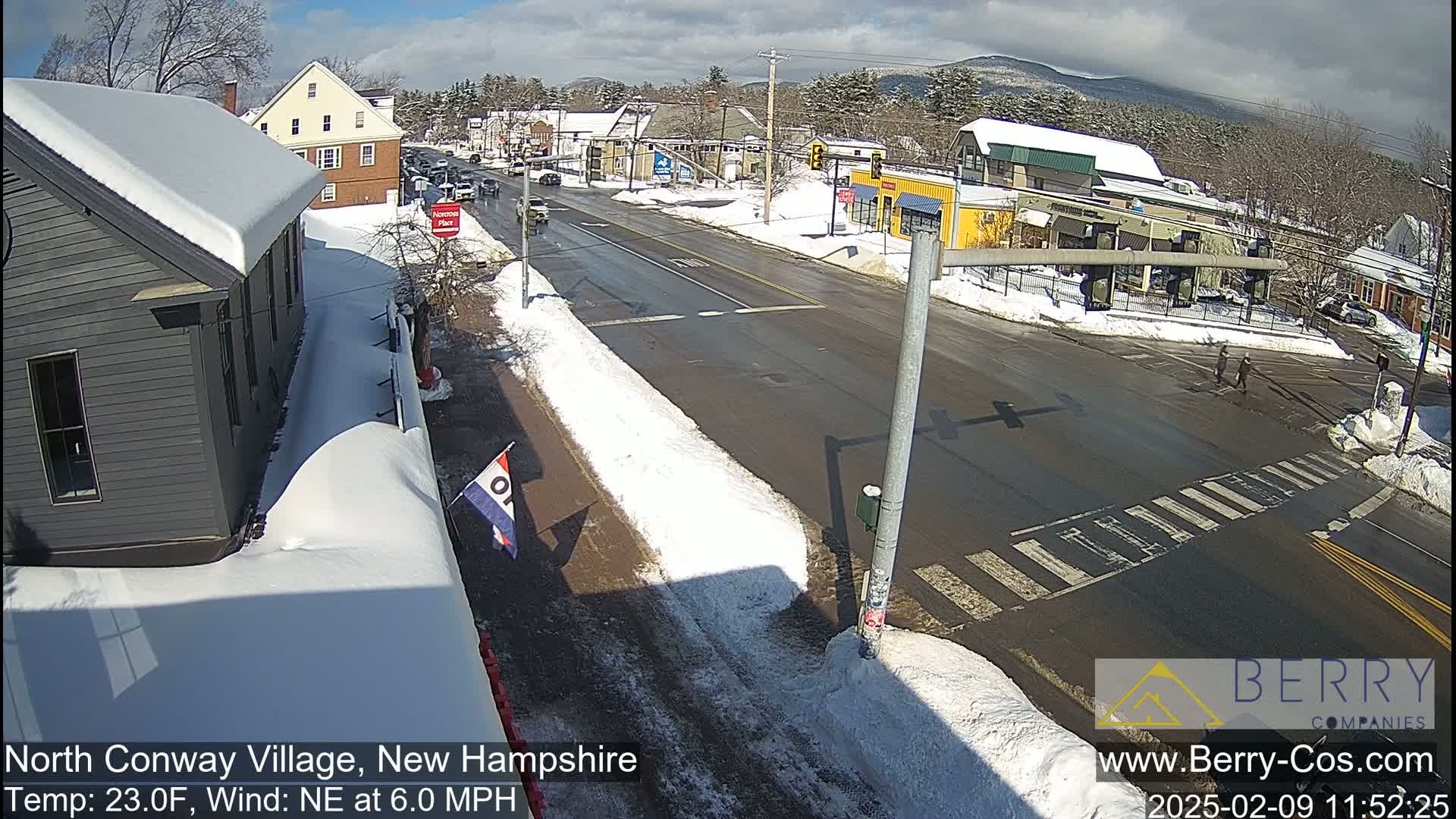 A snow-covered street scene in a village shows buildings, cars, and pedestrians on a sunny day with a northeast wind.