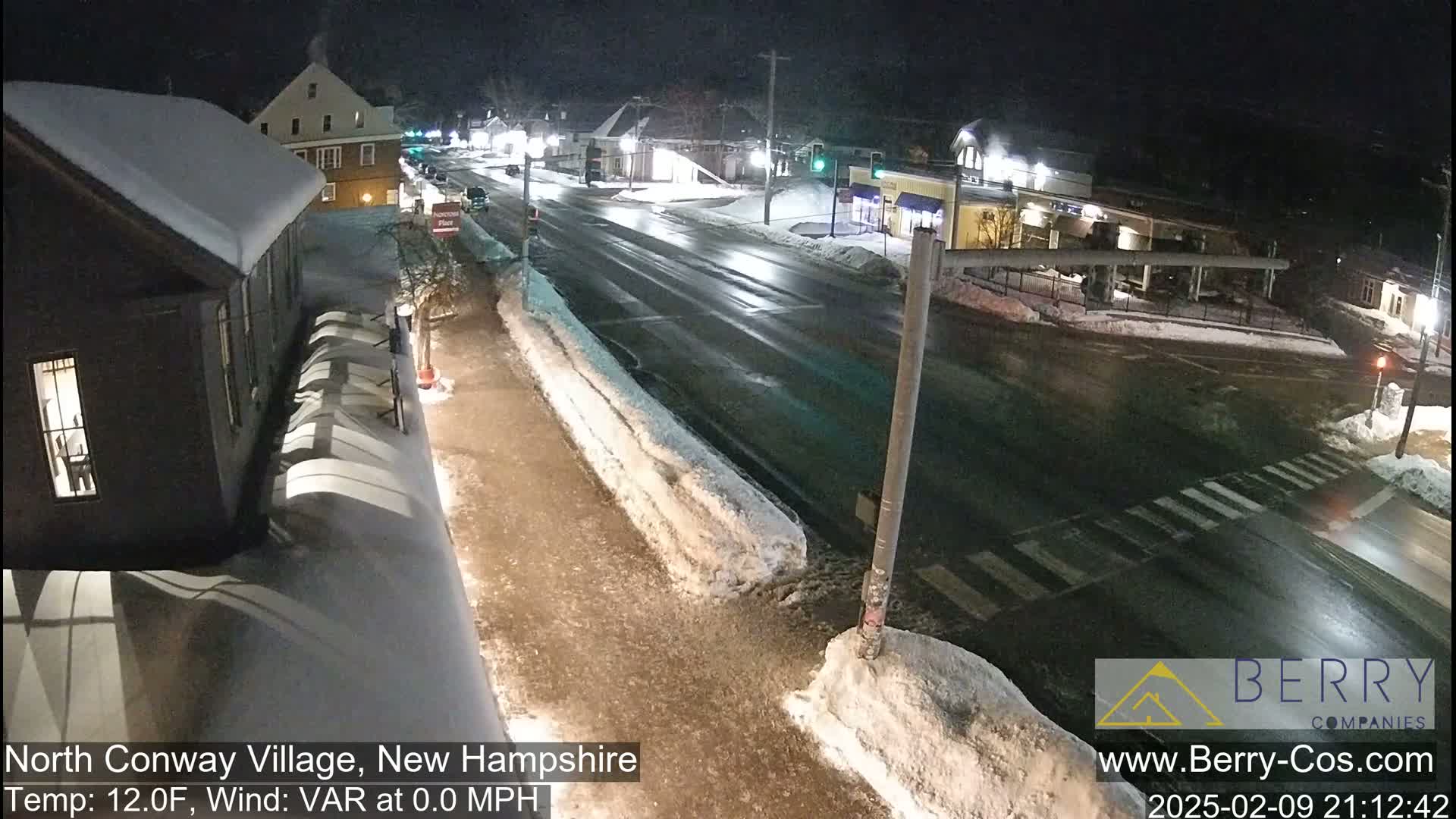 A snow-covered town street scene at night, with a few cars and buildings visible under dim lighting.