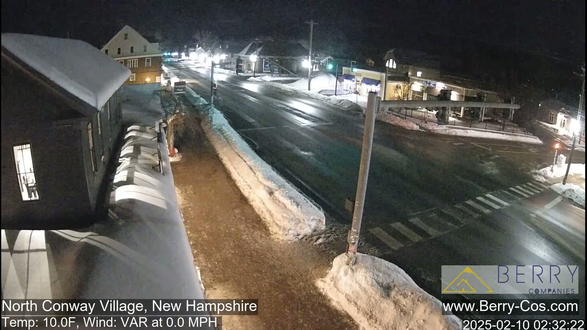 A snow-covered street scene at night shows a quiet intersection with buildings and minimal traffic under calm, clear conditions.