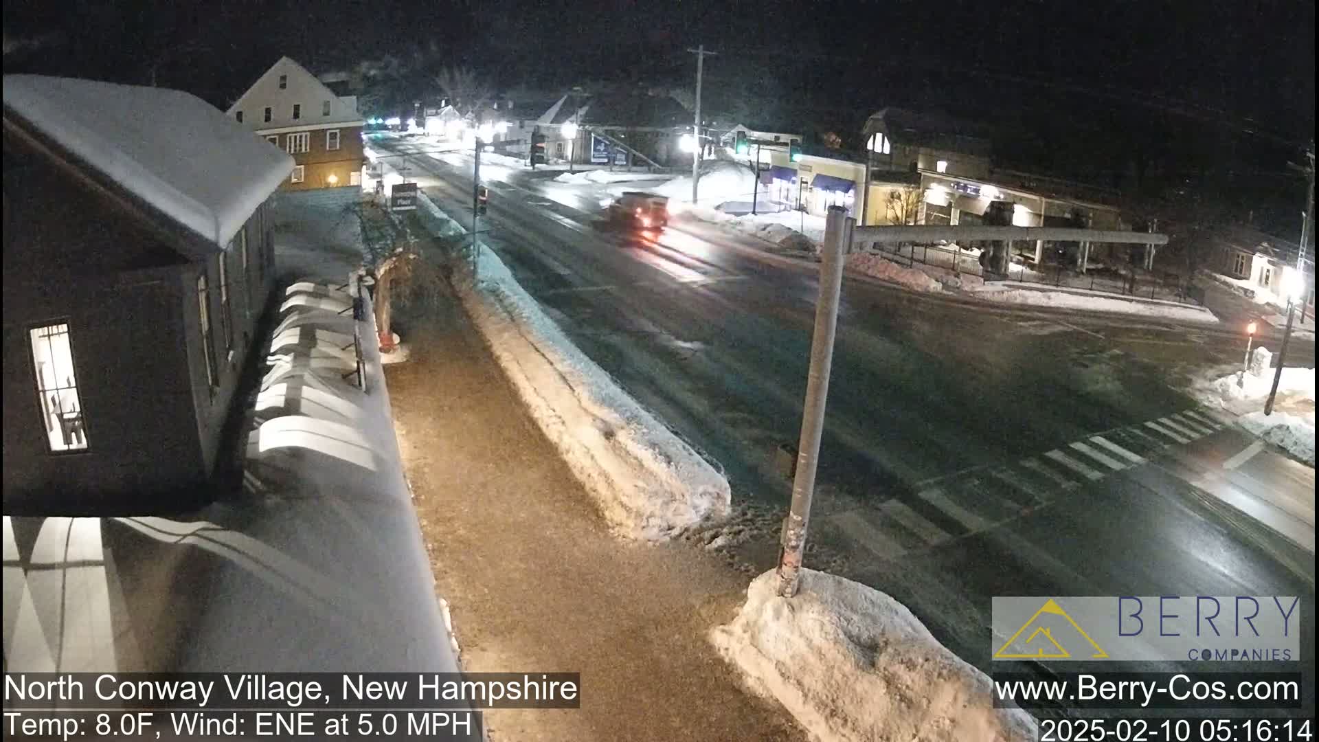 A snow-covered street scene at night shows a vehicle driving on a road near buildings, with a light dusting of snow on the ground and some snow piled along the curb.