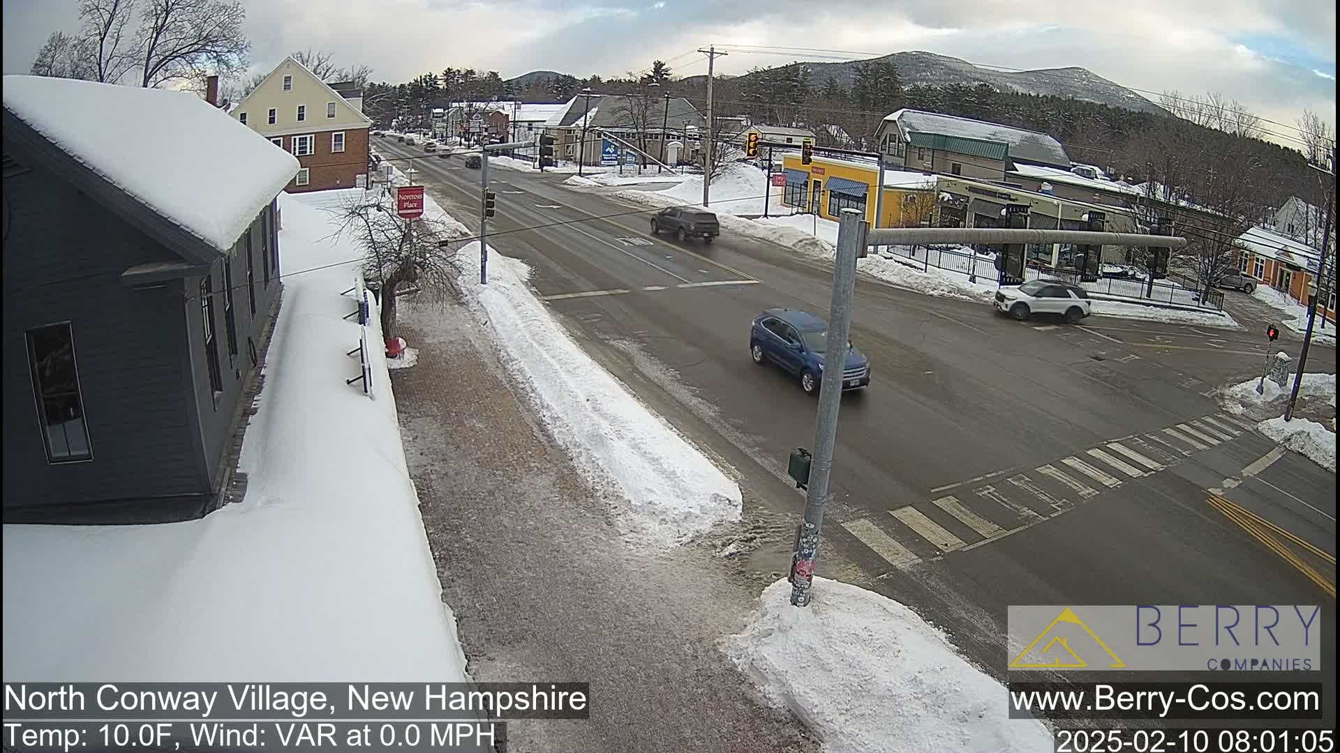 A snow-covered street scene in a small town shows a few cars at an intersection under a cloudy sky.