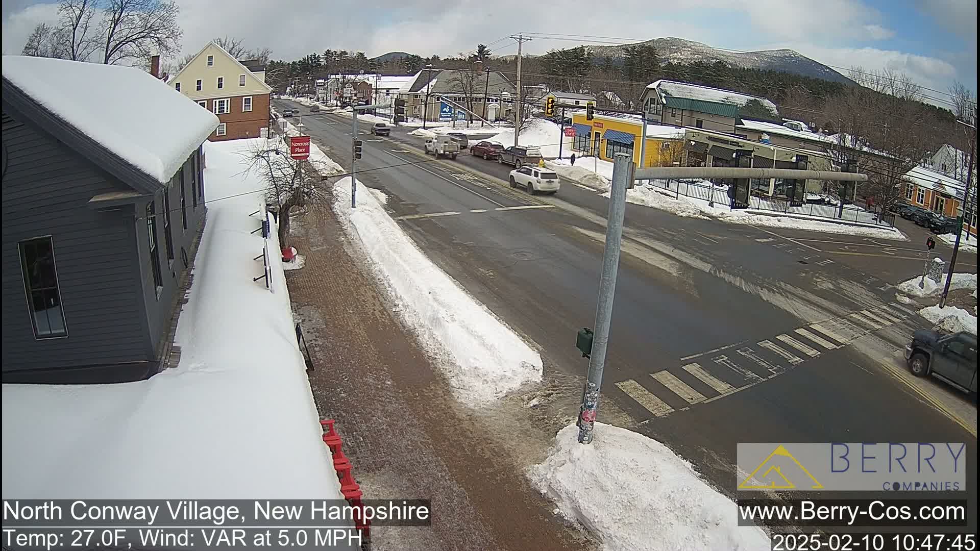 A snow-covered street scene in a small town shows several cars at an intersection, with mountains visible in the background under a partly cloudy sky.