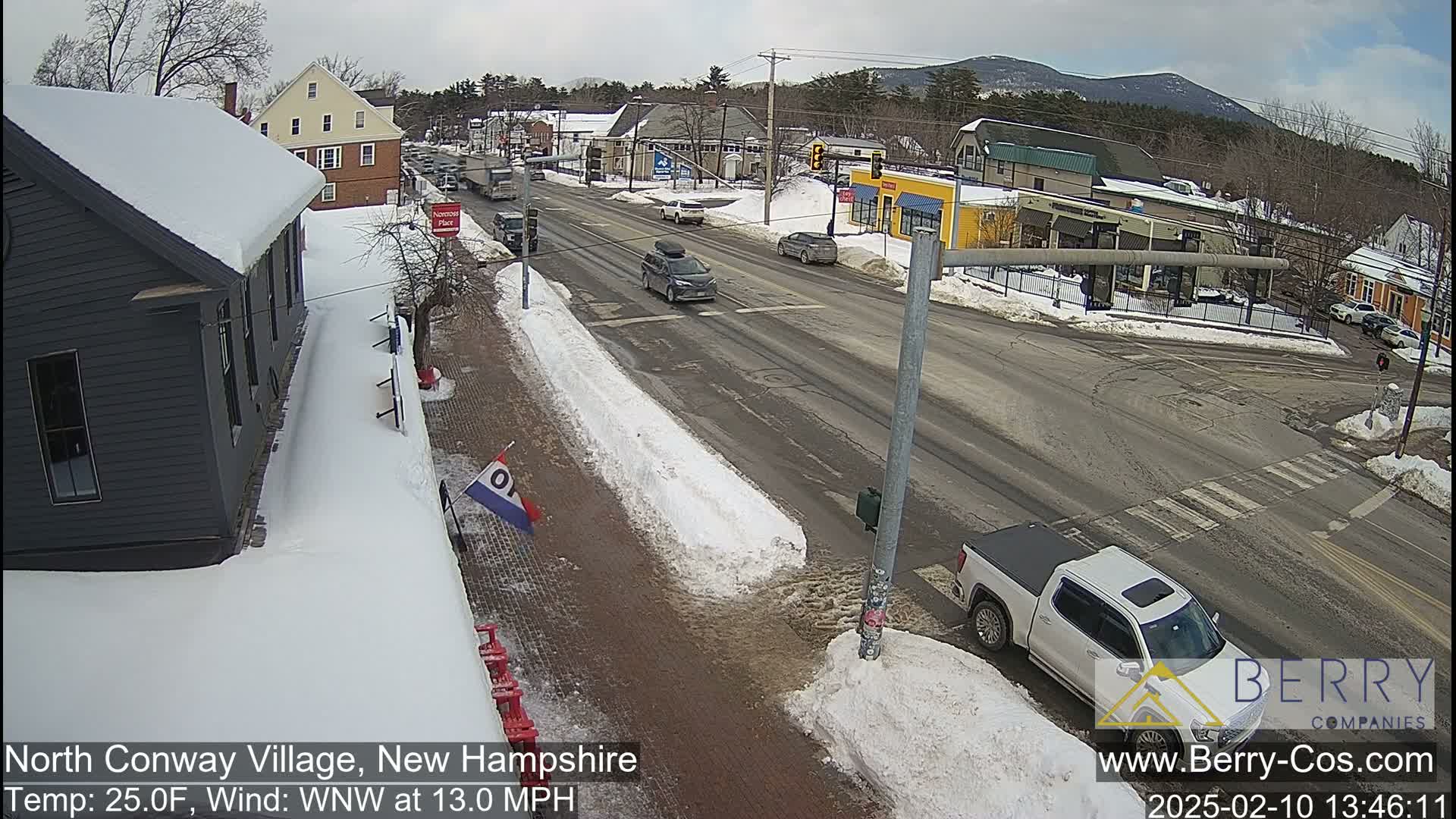 A snow-covered street scene shows several cars driving on a road with buildings and businesses on either side, under a partly cloudy sky.