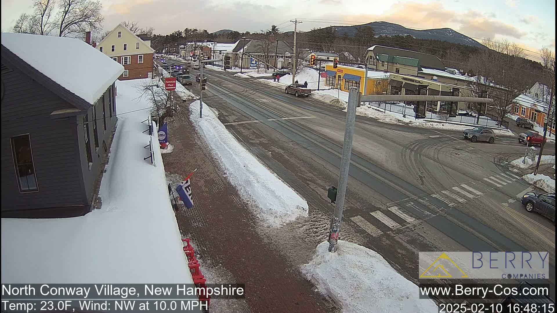 A snow-covered street scene shows a town intersection with several cars, buildings, and snow-covered sidewalks under a partly cloudy sky.