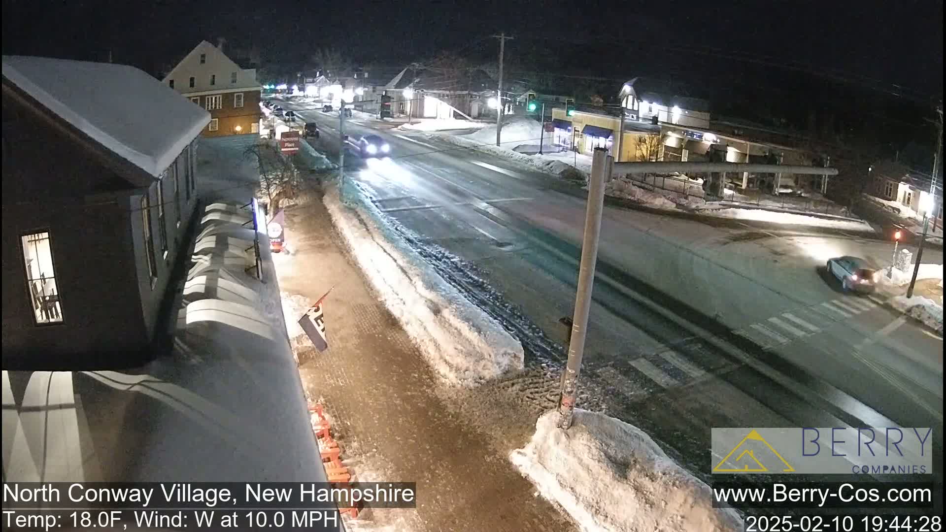 A snow-covered street scene at night shows a few cars driving on a road lined with buildings, under a clear sky.