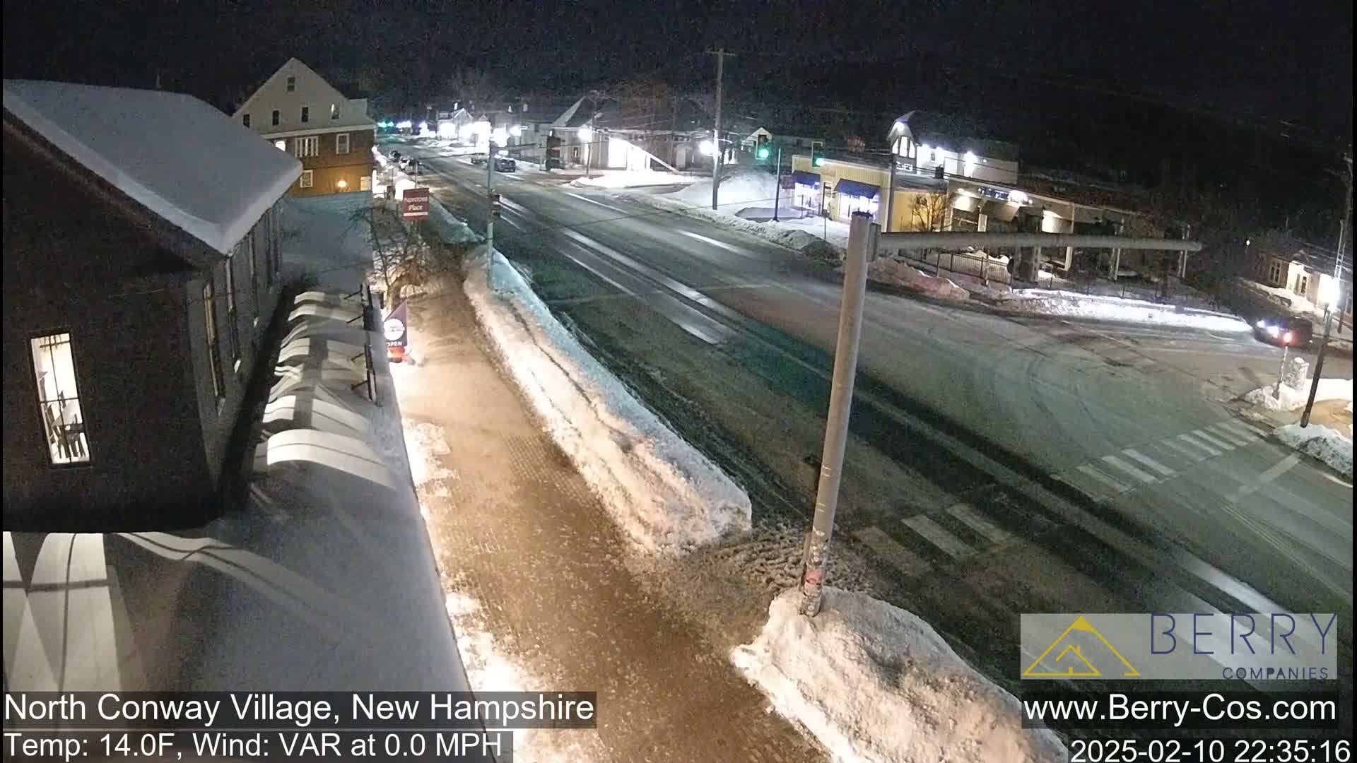 A snow-covered street scene at night shows a road intersection with buildings and sidewalks lined with snow.