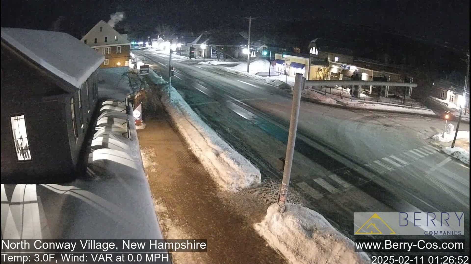 A snow-covered street scene at night, showing buildings and a road intersection with light snowfall.