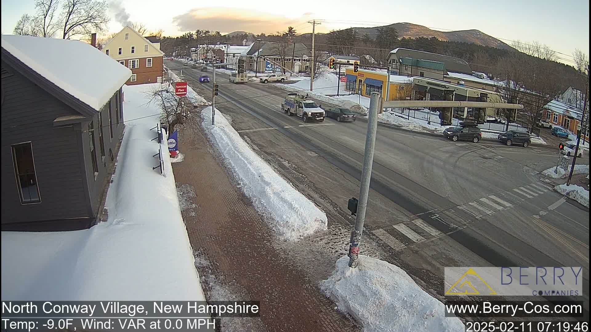 A snow-covered street scene shows several vehicles at an intersection, with buildings and snow-covered sidewalks on a partly cloudy day.