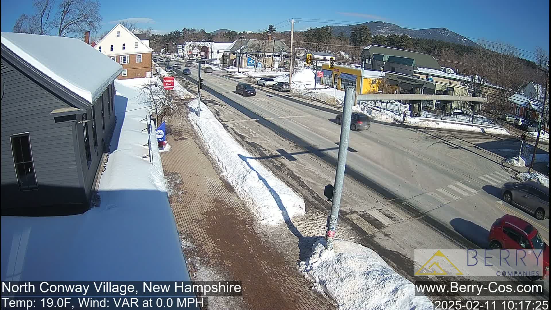 A sunny winter scene shows a snow-covered street with cars driving past buildings in a village.