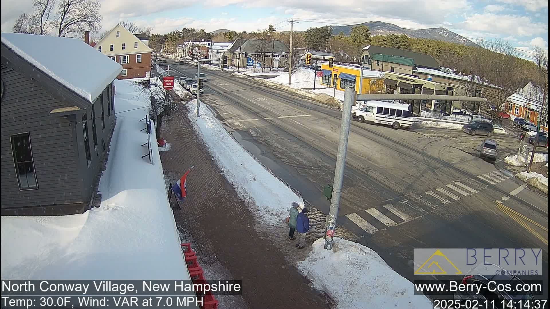 A snow-covered street scene shows a village intersection with cars and pedestrians, under a partly cloudy sky.