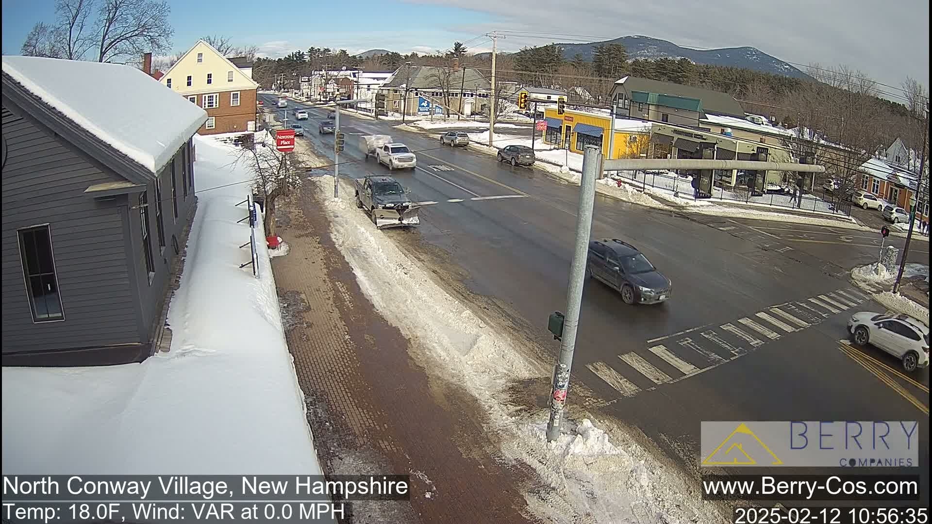 A snow-covered street scene shows a village intersection with several cars and a snowplow, under a sunny sky.