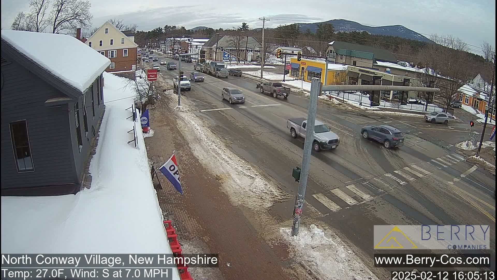 A snow-covered street scene shows several vehicles driving on a road in front of buildings on a partly sunny day with a south wind.