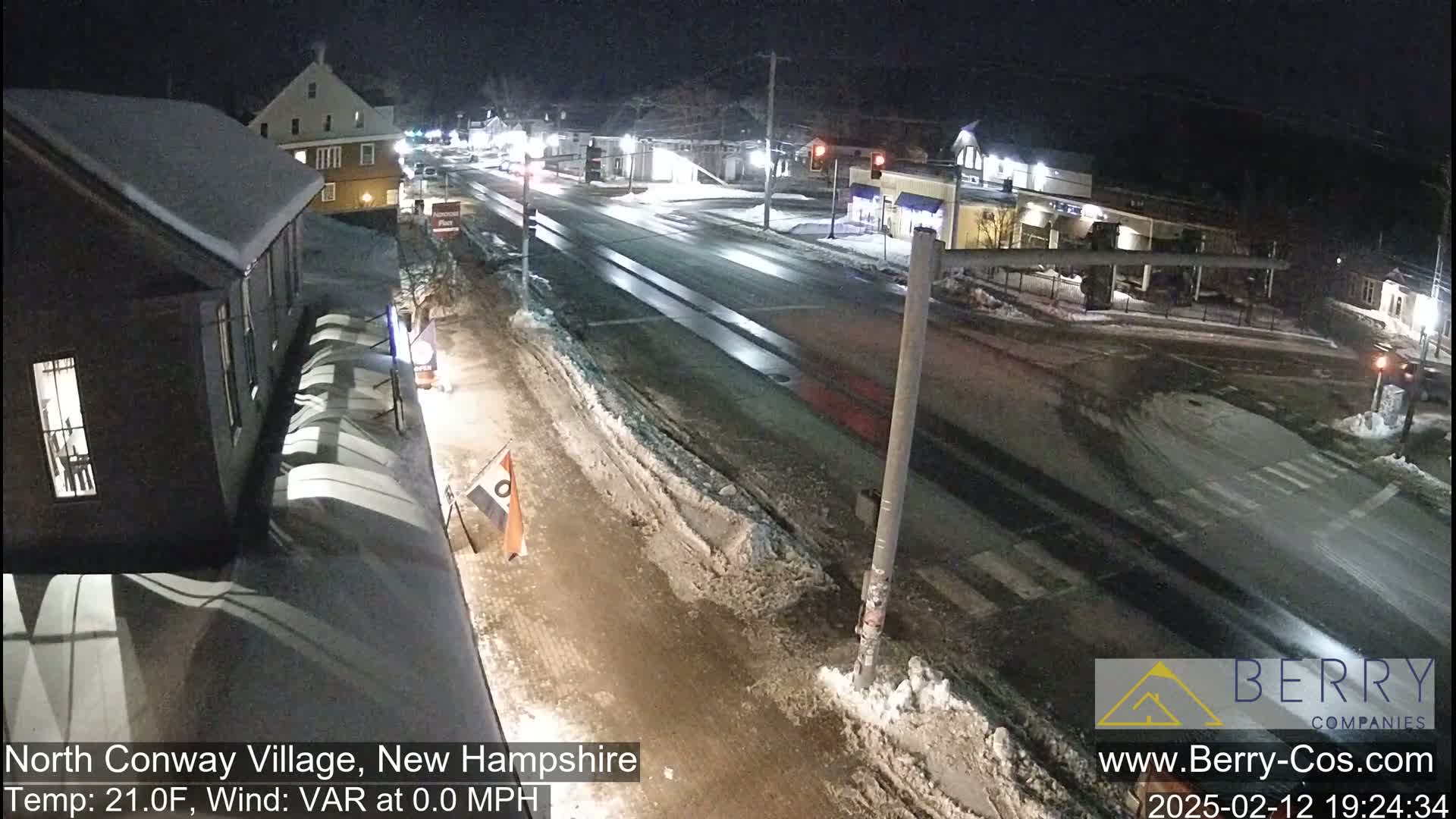A snow-covered street scene at night, showing a multi-lane road with buildings and some traffic.