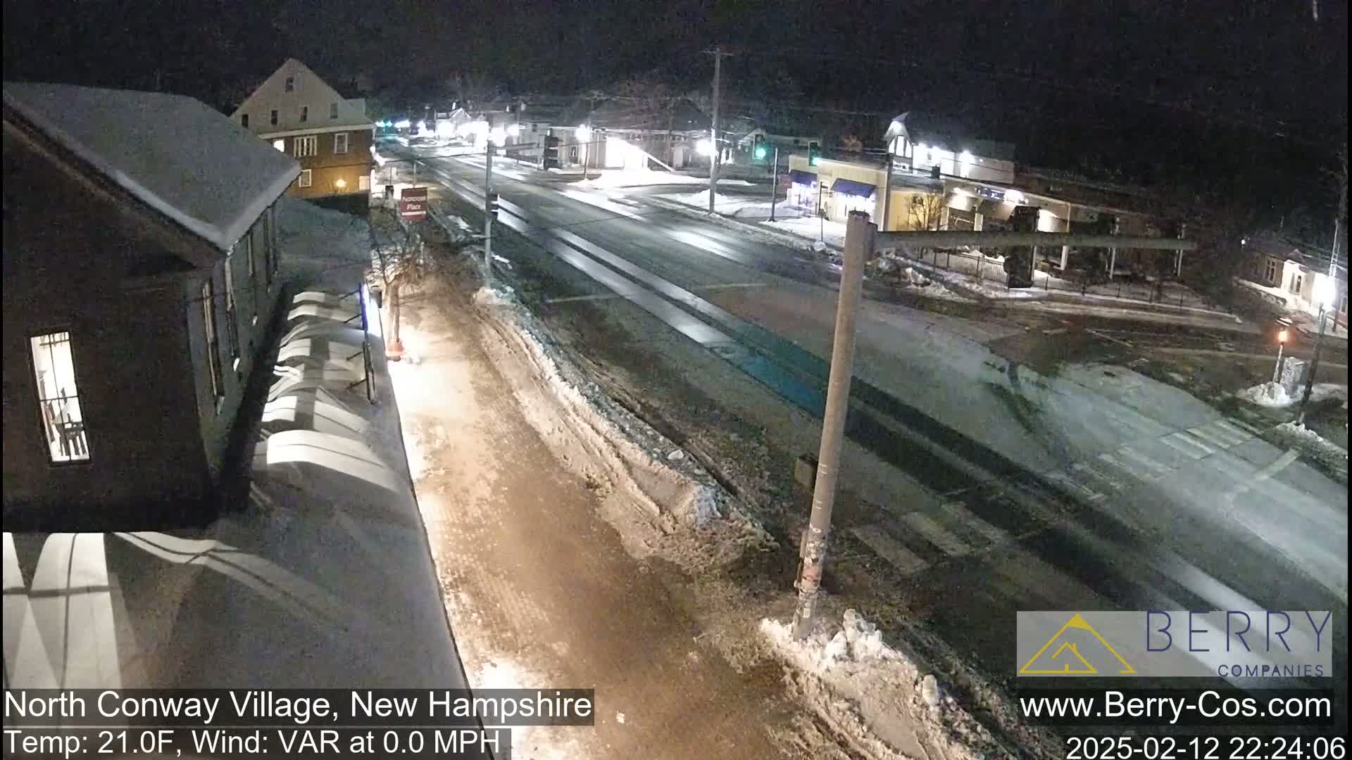 A snow-covered street scene at night shows a mostly empty road with buildings and businesses on either side.