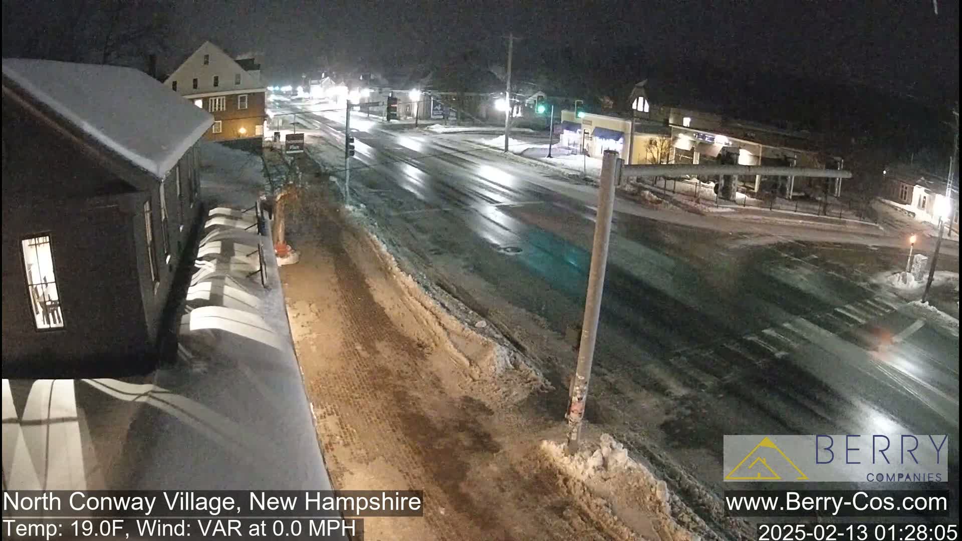 A snow-covered street scene at night, with buildings visible on either side and a few cars on the road.