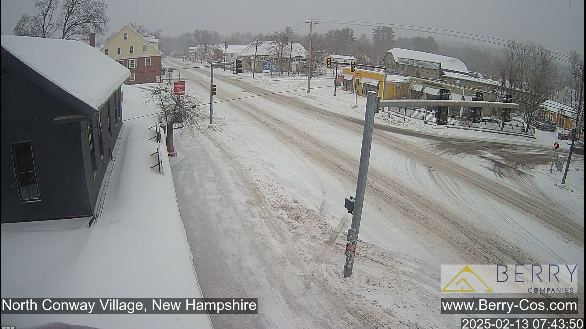 A snow-covered street intersection in a small town, with several buildings visible and light snowfall.