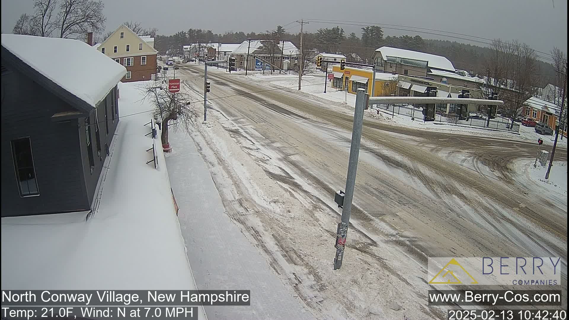 A snow-covered street scene shows a road intersection in a small town, with buildings on either side, under an overcast sky.