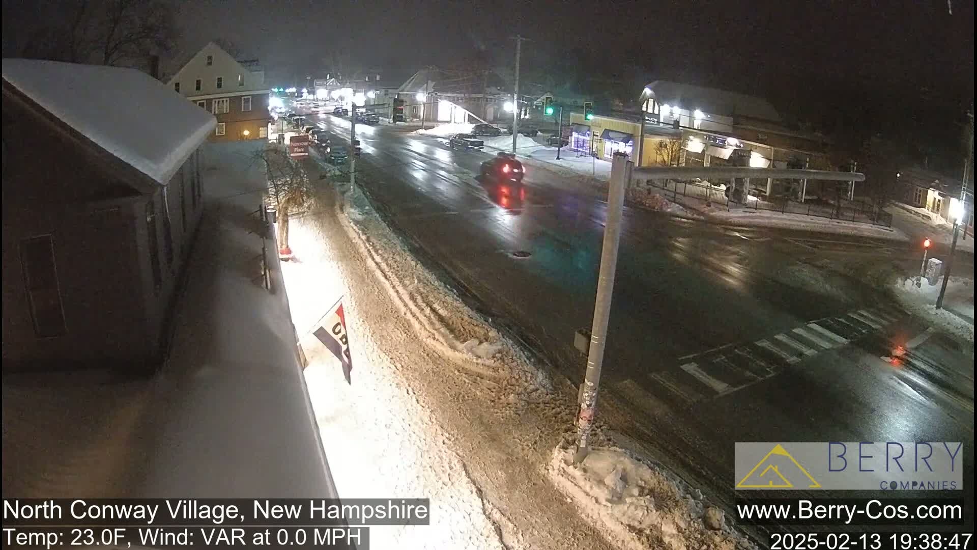 A snow-covered street intersection at night with a few vehicles and buildings illuminated by streetlights.