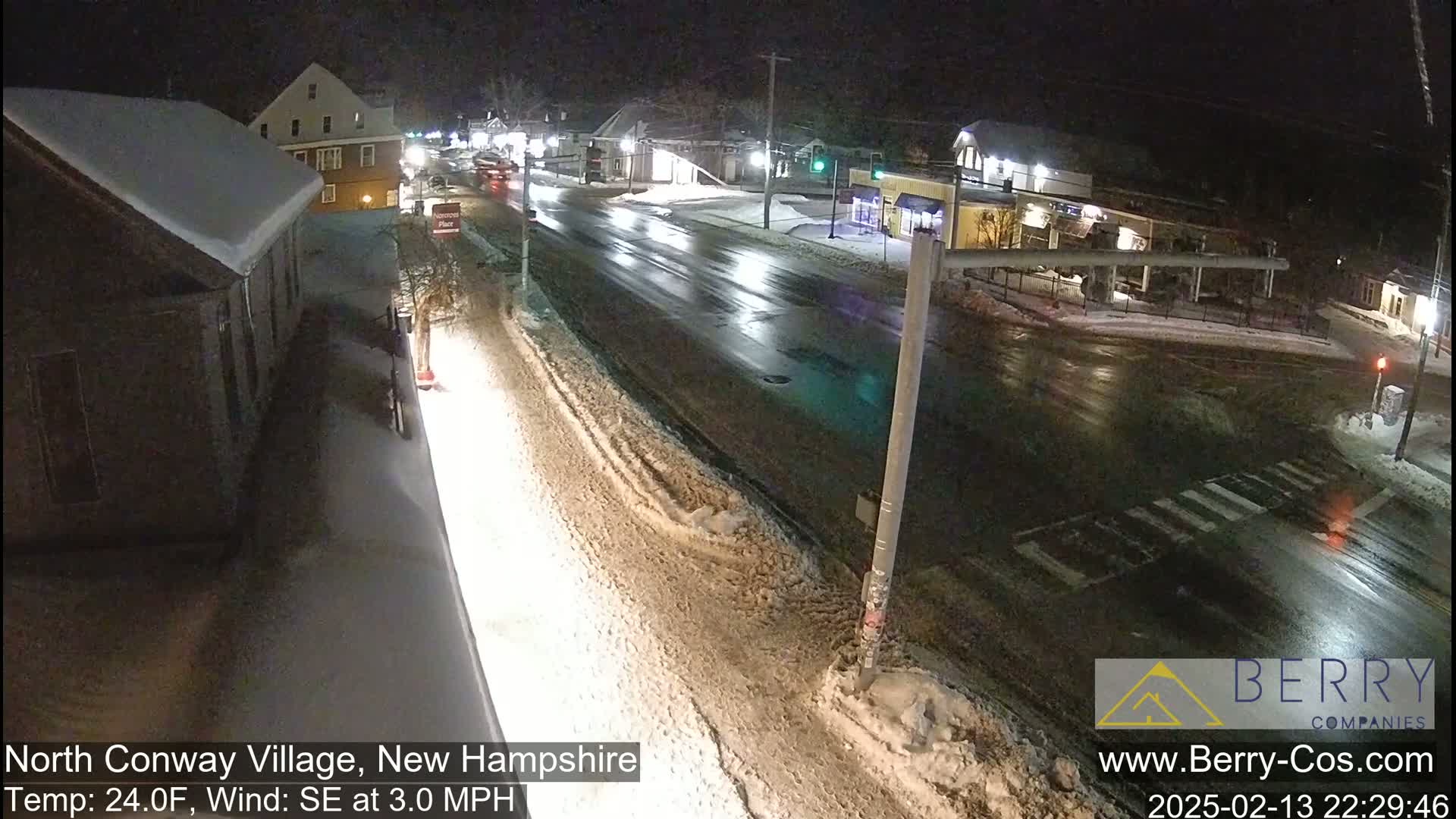 A snow-covered street scene at night shows a roadway intersection with a few vehicles and buildings under a dark sky.
