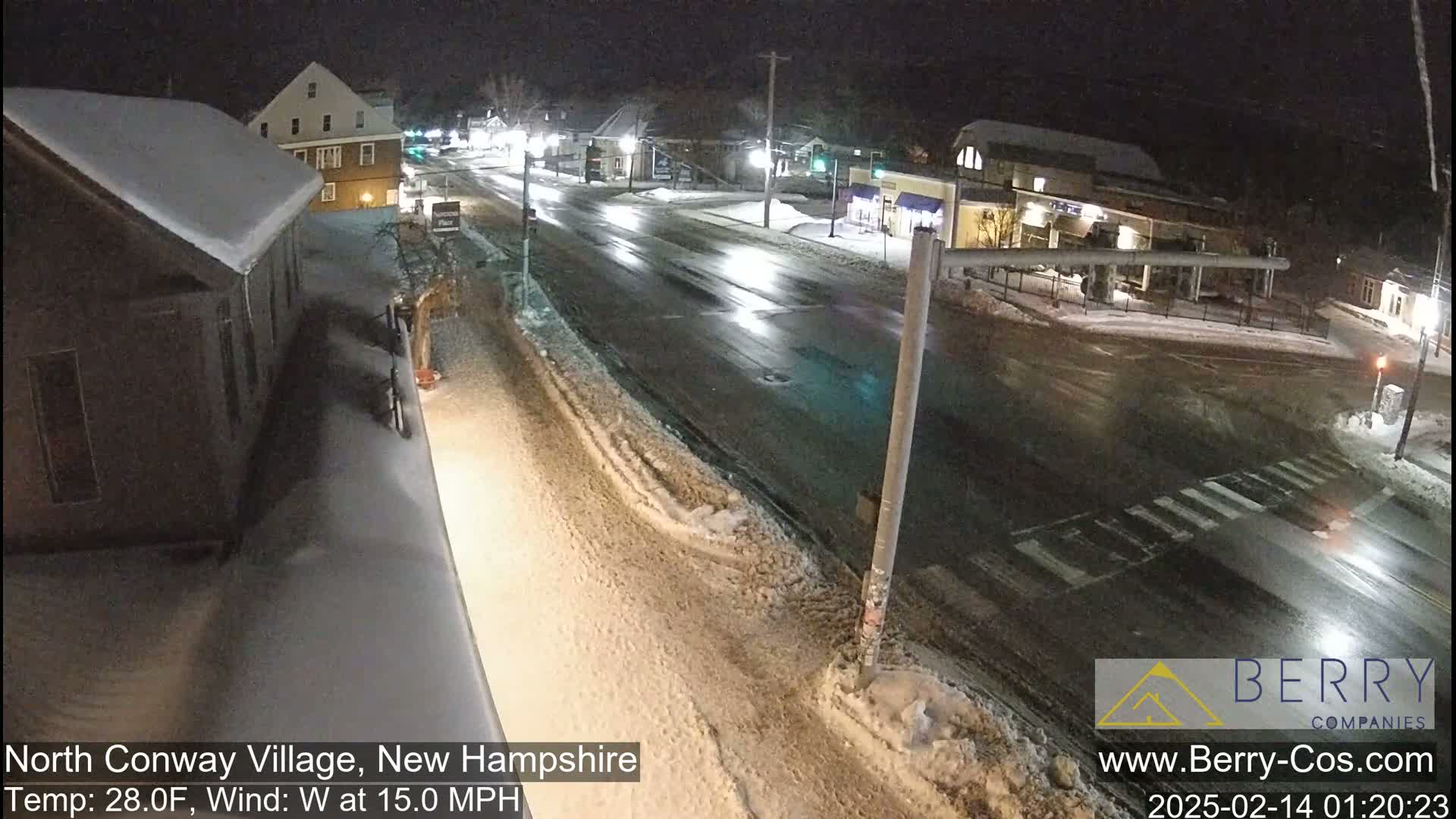 A snow-covered street scene at night, with buildings and cars visible under a partly cloudy sky and a 15 mph west wind.