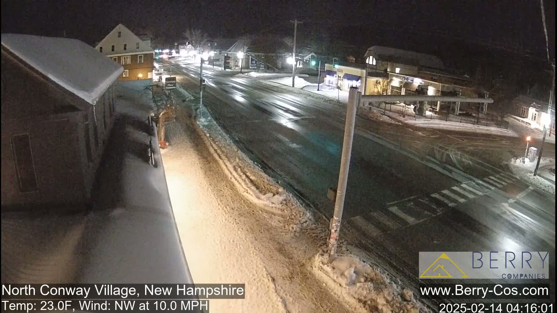 A snow-covered street scene at night shows a road intersection with buildings on either side, under a nighttime sky with a northwest wind blowing at 10 mph and a temperature of 23 degrees Fahrenheit.