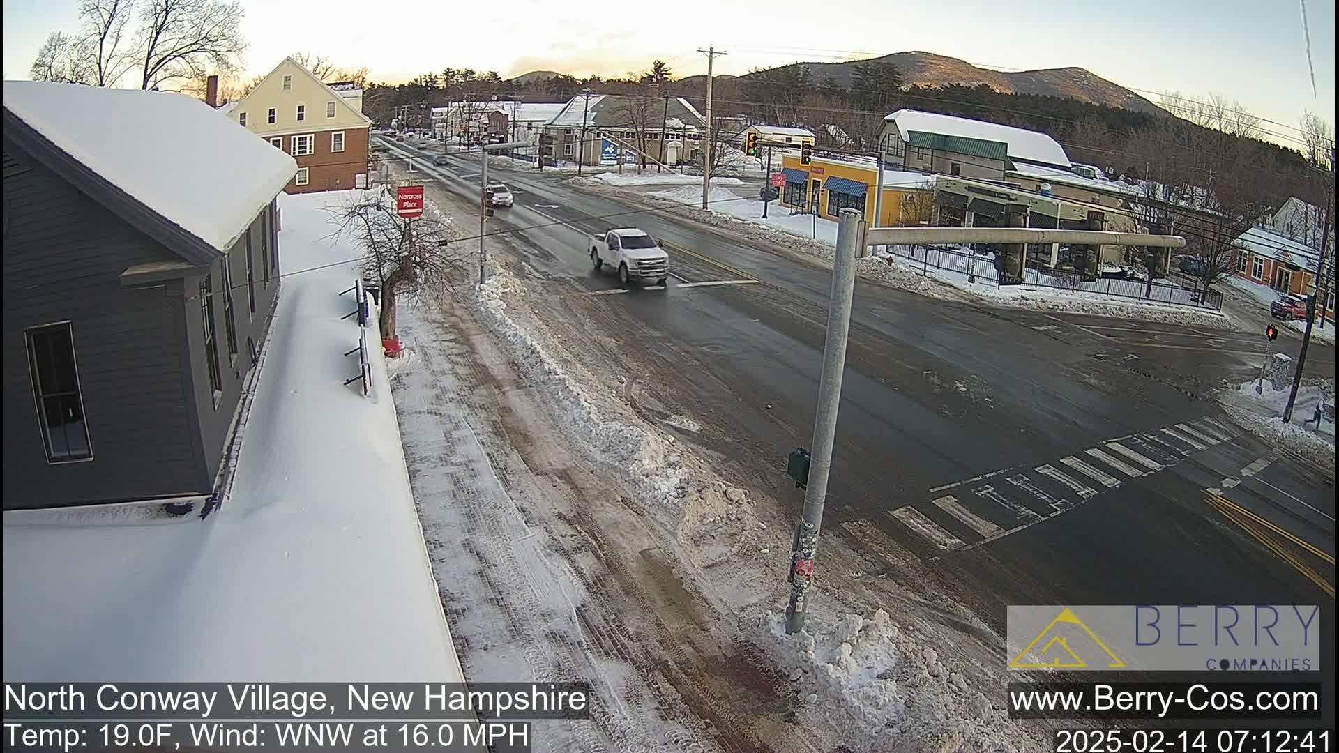 A snow-covered street scene shows a crossroads in a small town with a few vehicles, under a partly cloudy sky.