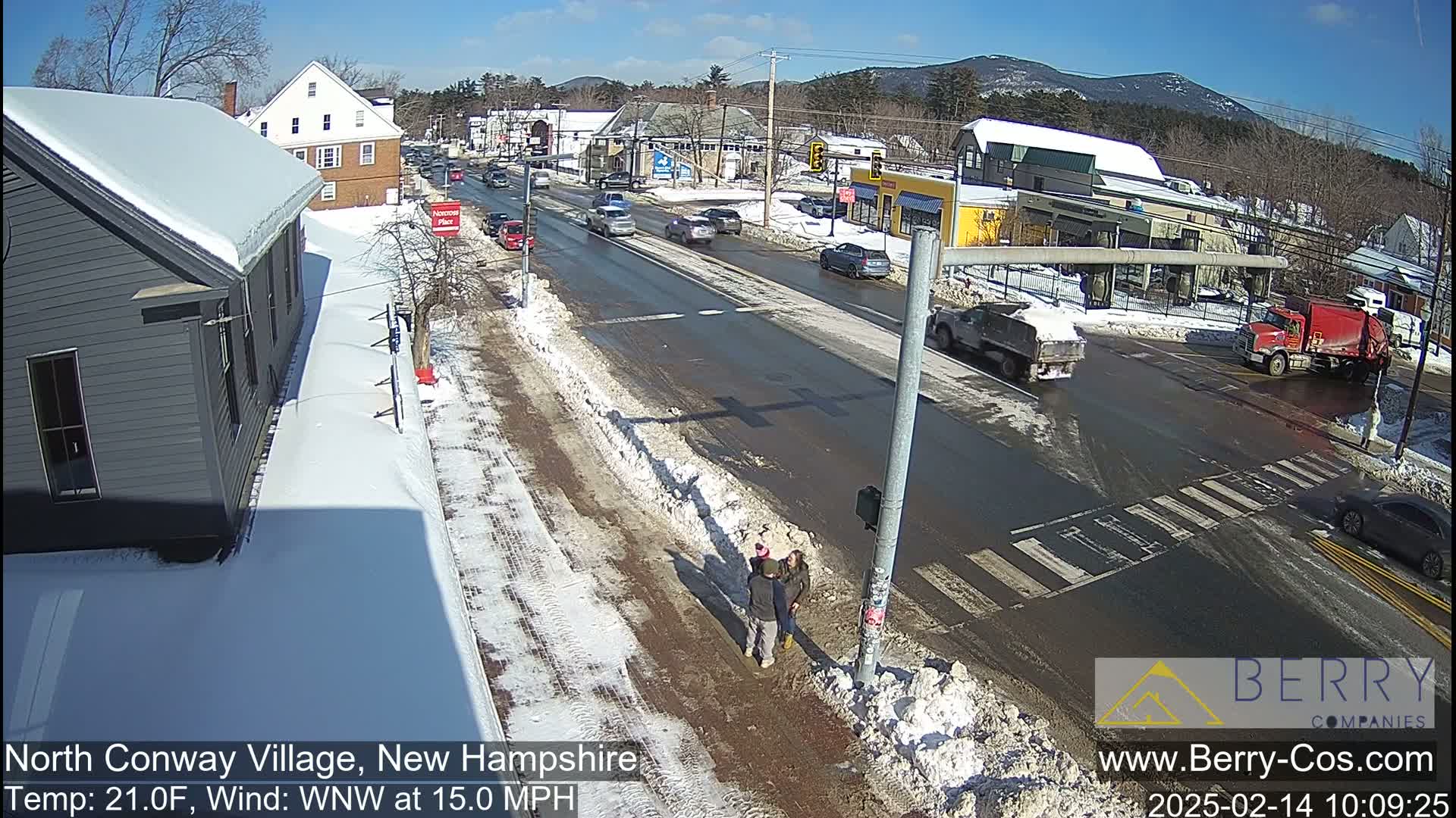 A sunny, snowy street scene shows a two-lane road with cars and trucks, pedestrians at a crosswalk, and snow-covered sidewalks and buildings.