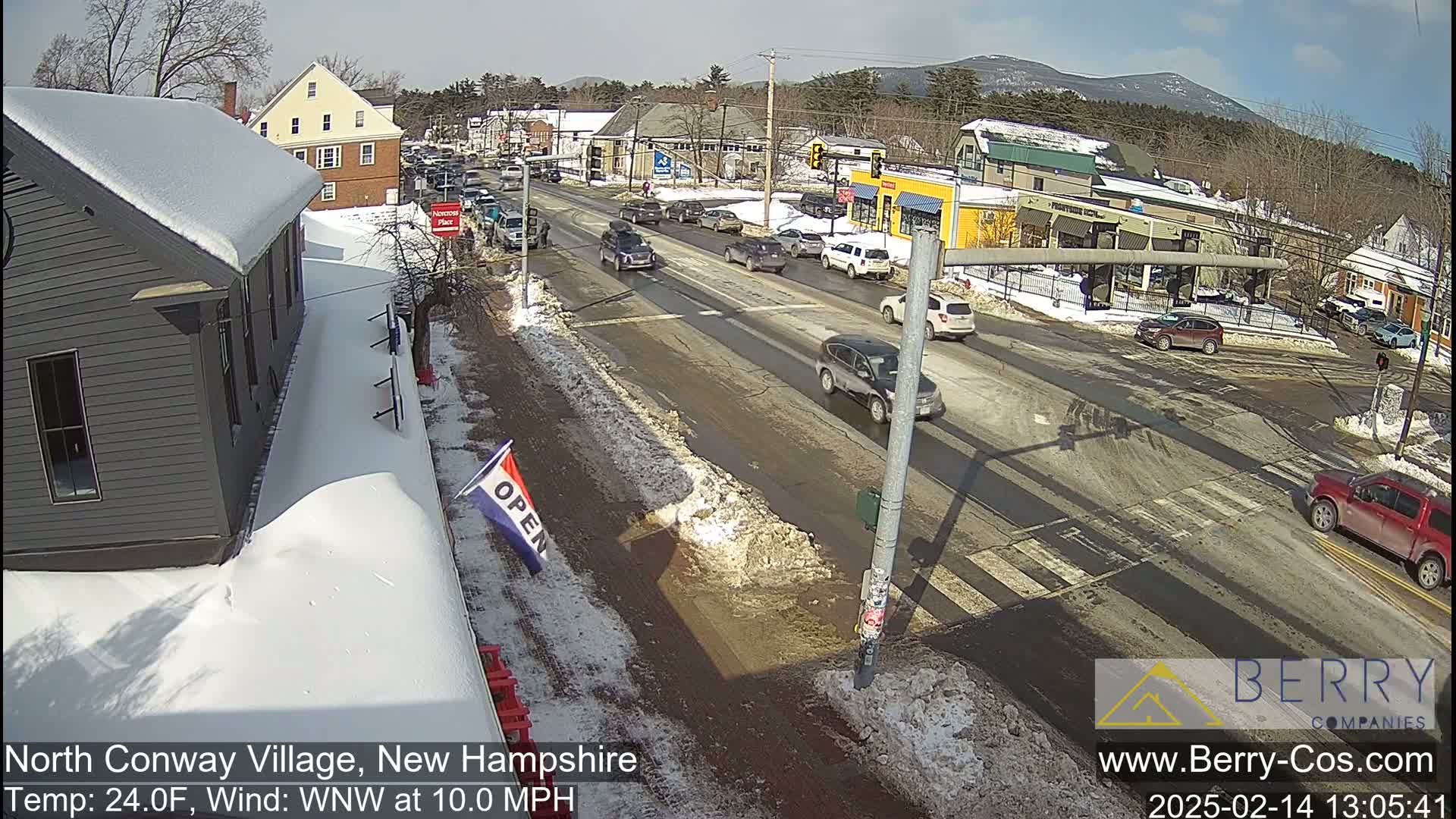 A sunny day with snow on the ground shows a village street with cars driving through an intersection and buildings lining the road.