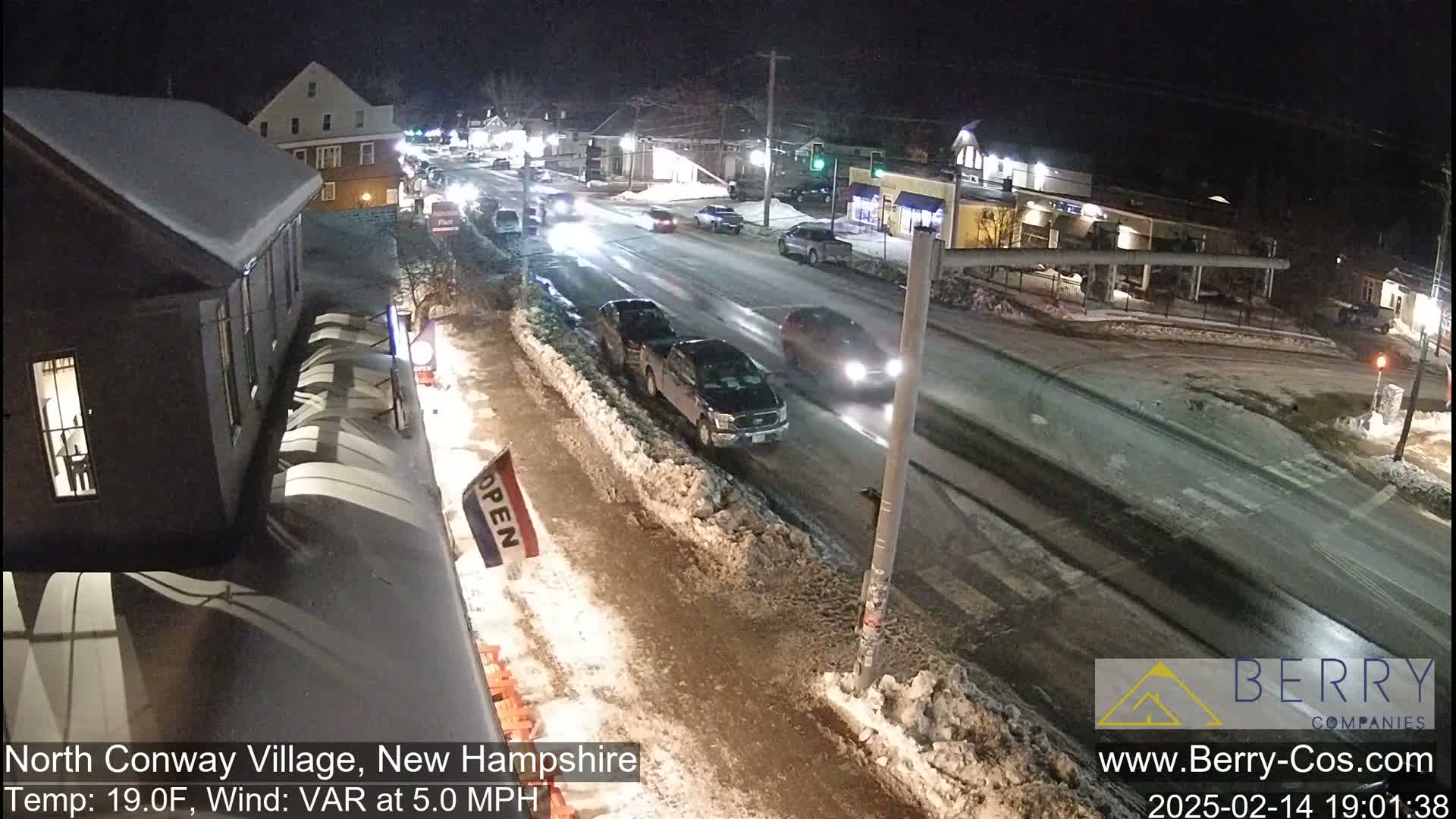 A snow-covered street at night with several cars driving on it,  viewed from a slightly elevated position, showing a partly snow-covered sidewalk and buildings.
