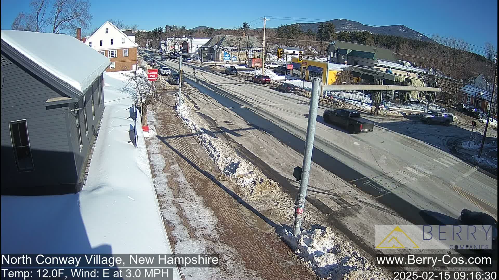 A sunny winter scene shows a snow-covered street with several cars and buildings, mountains in the background.