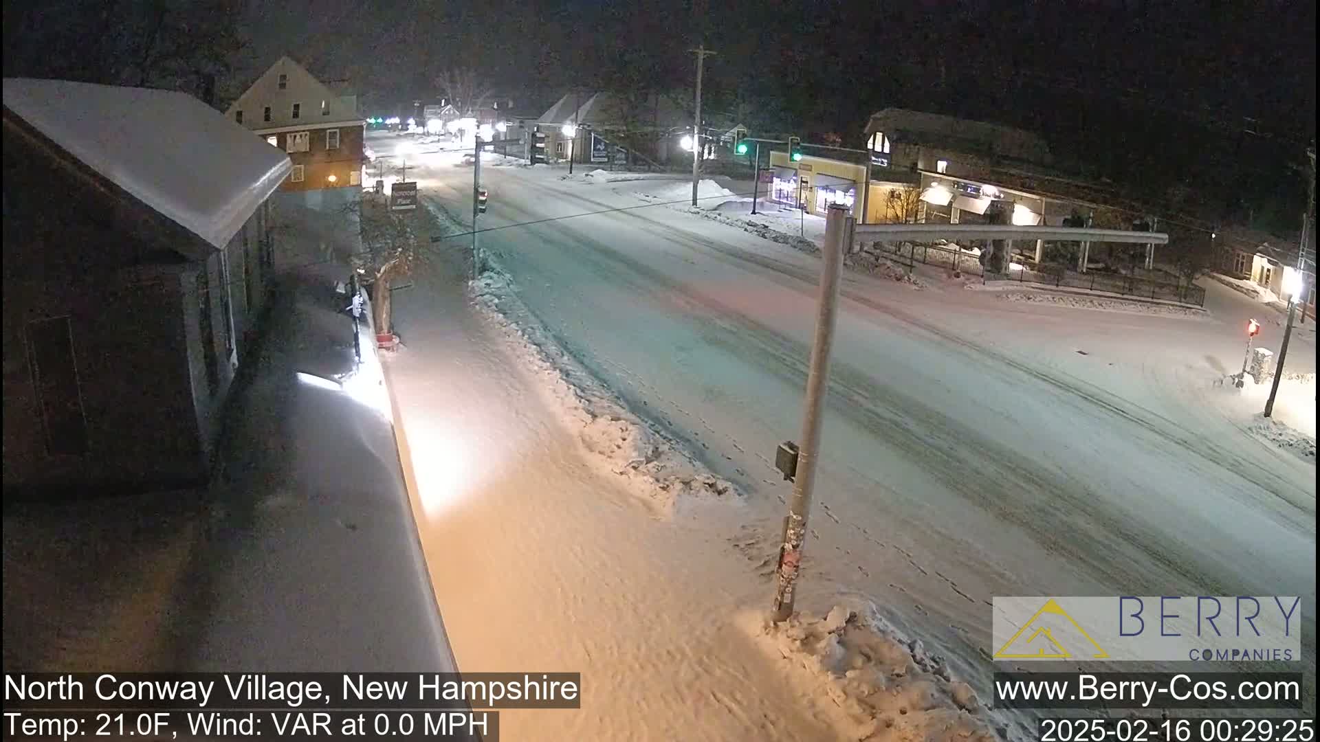 A snow-covered street scene at night shows a road with buildings and some cars, with a temperature of 21 degrees Fahrenheit and no wind.