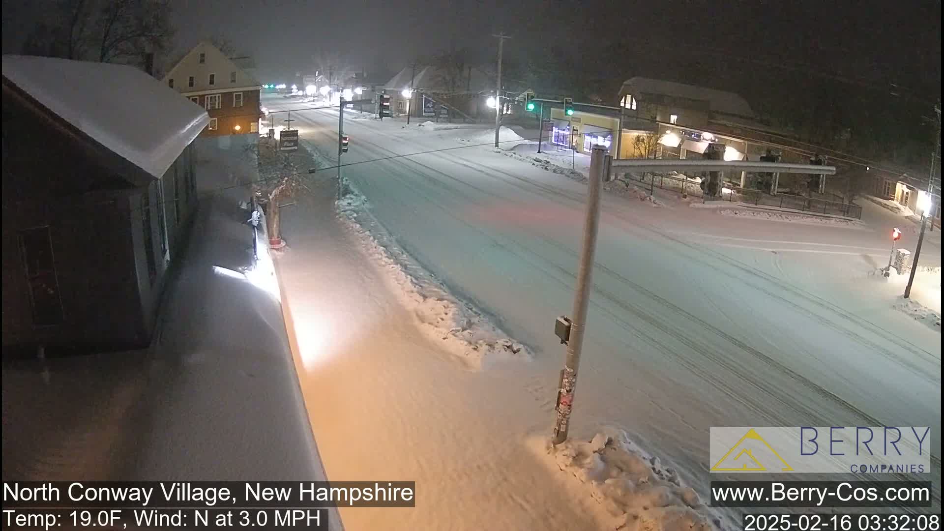 A snow-covered street scene at night, with buildings and streetlights visible under a light snowfall.