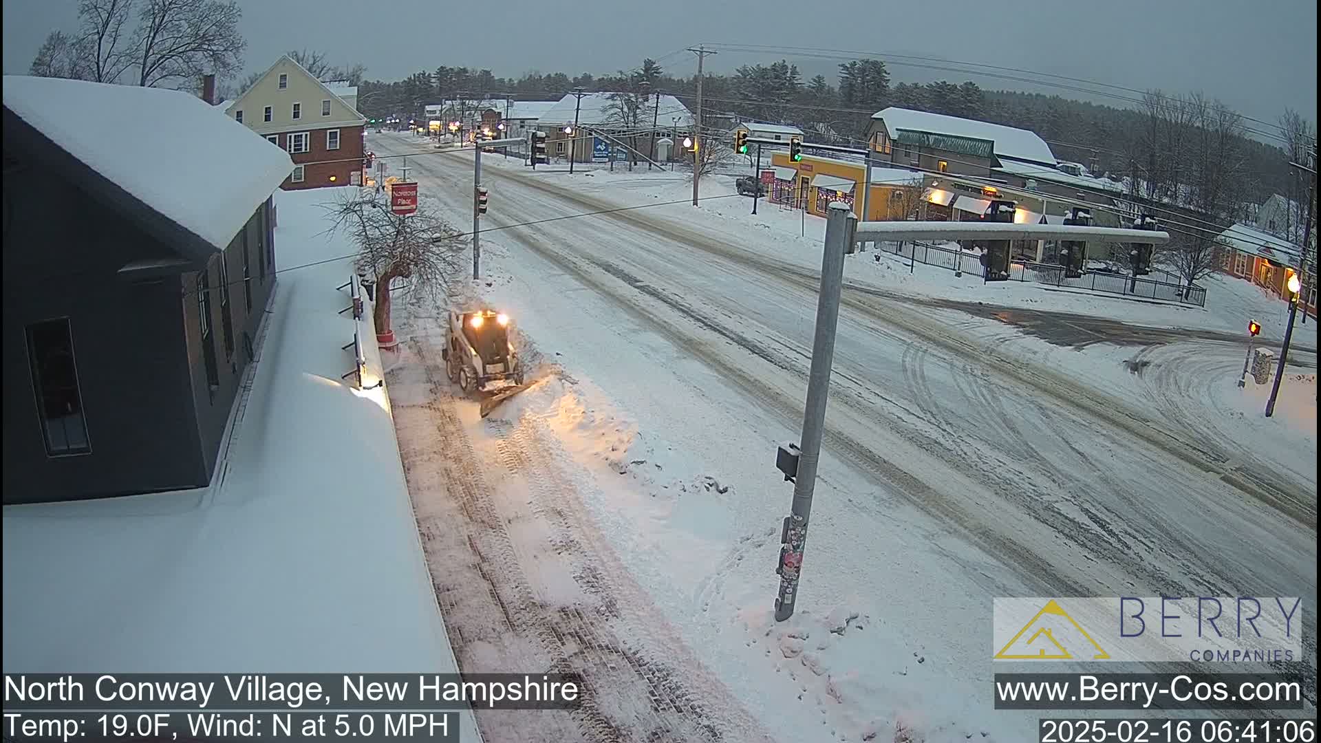 A snow plow clears a snow-covered street in a village, under a gray, overcast sky.